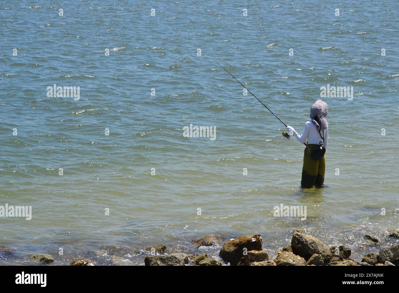 A fisherwoman, knee-deep in the ocean, holds a fishing rod as she looks ...