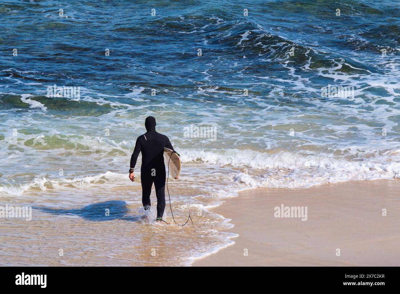 A surfer wearing a full body wetsuit carrying a surfboard heads into