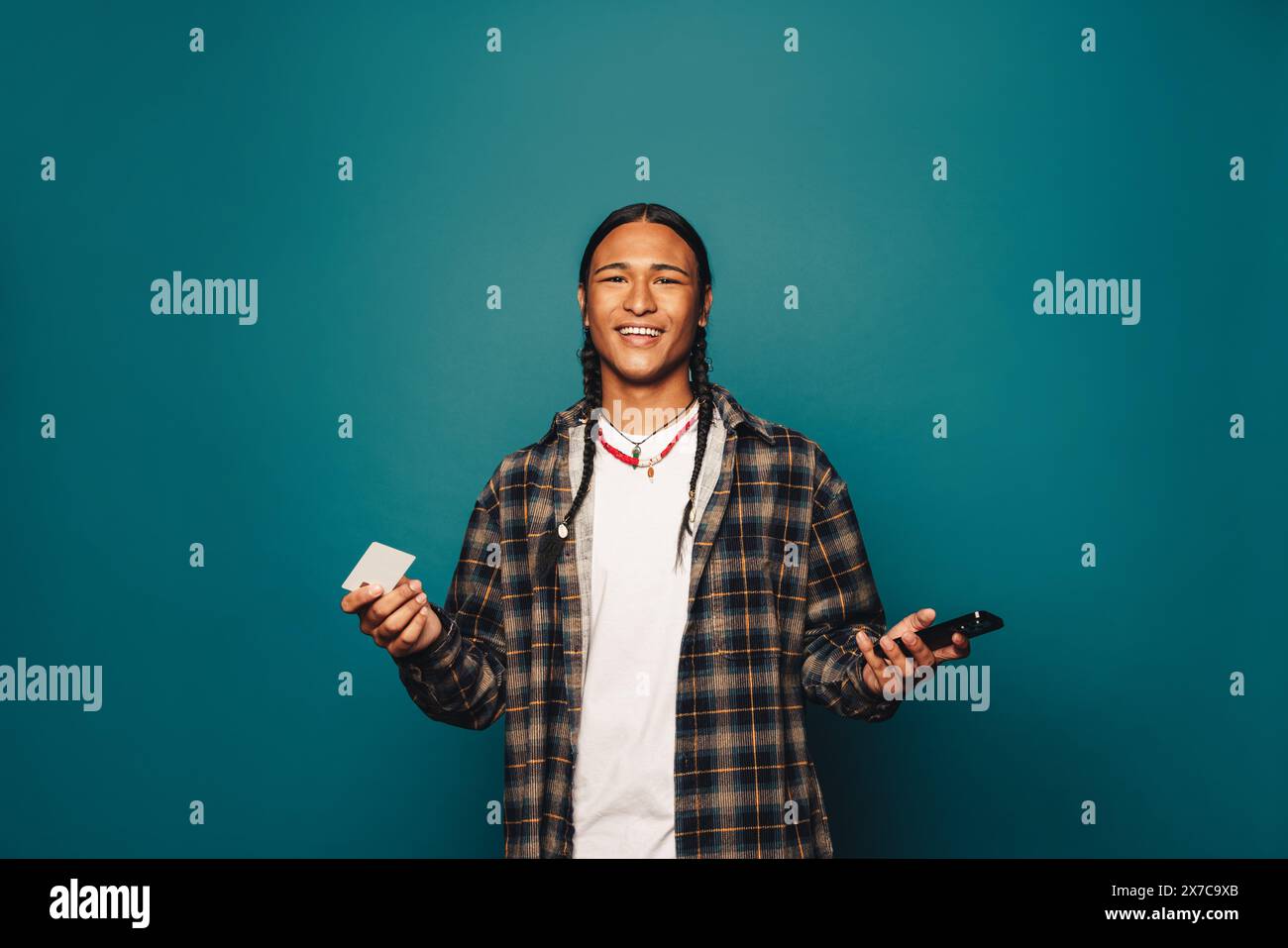 Cheerful, stylish person with braided hair stands against a blue background, holding a premium credit card and cellphone. Man representing the conveni Stock Photo