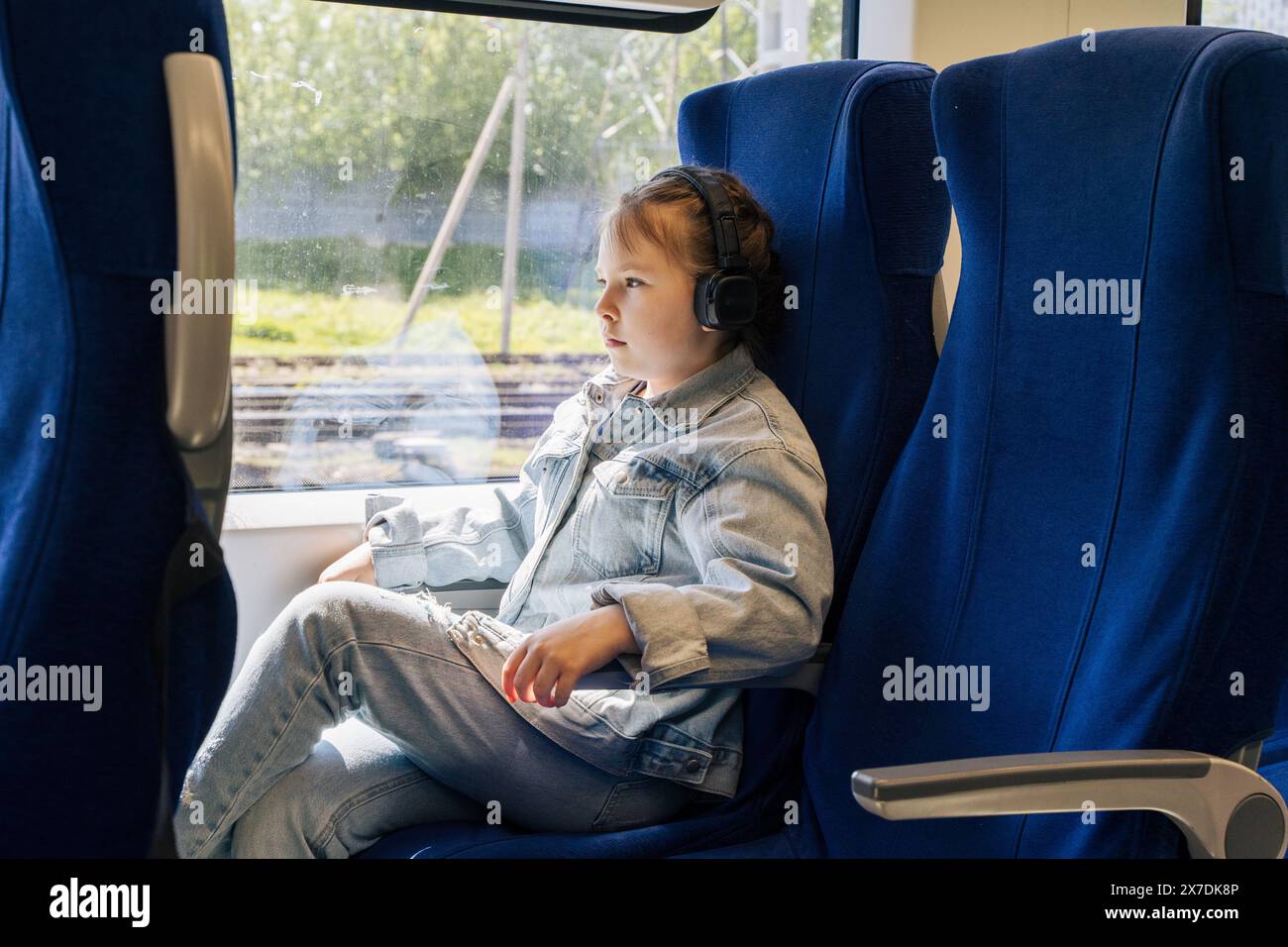 A little girl is traveling by train alone. A girl in headphones is sitting in a blue chair on the train and listening to music. pleasure in traveling, Stock Photo