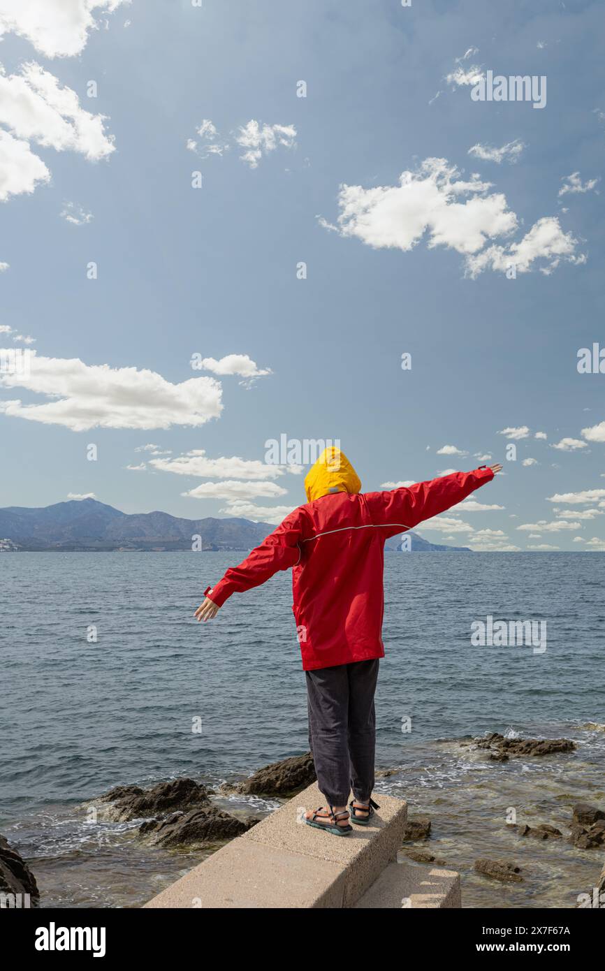 person in front of the sea with a storm overhead with large gray clouds, weather change red jacket with yellow cap, arms raised in protest Stock Photo