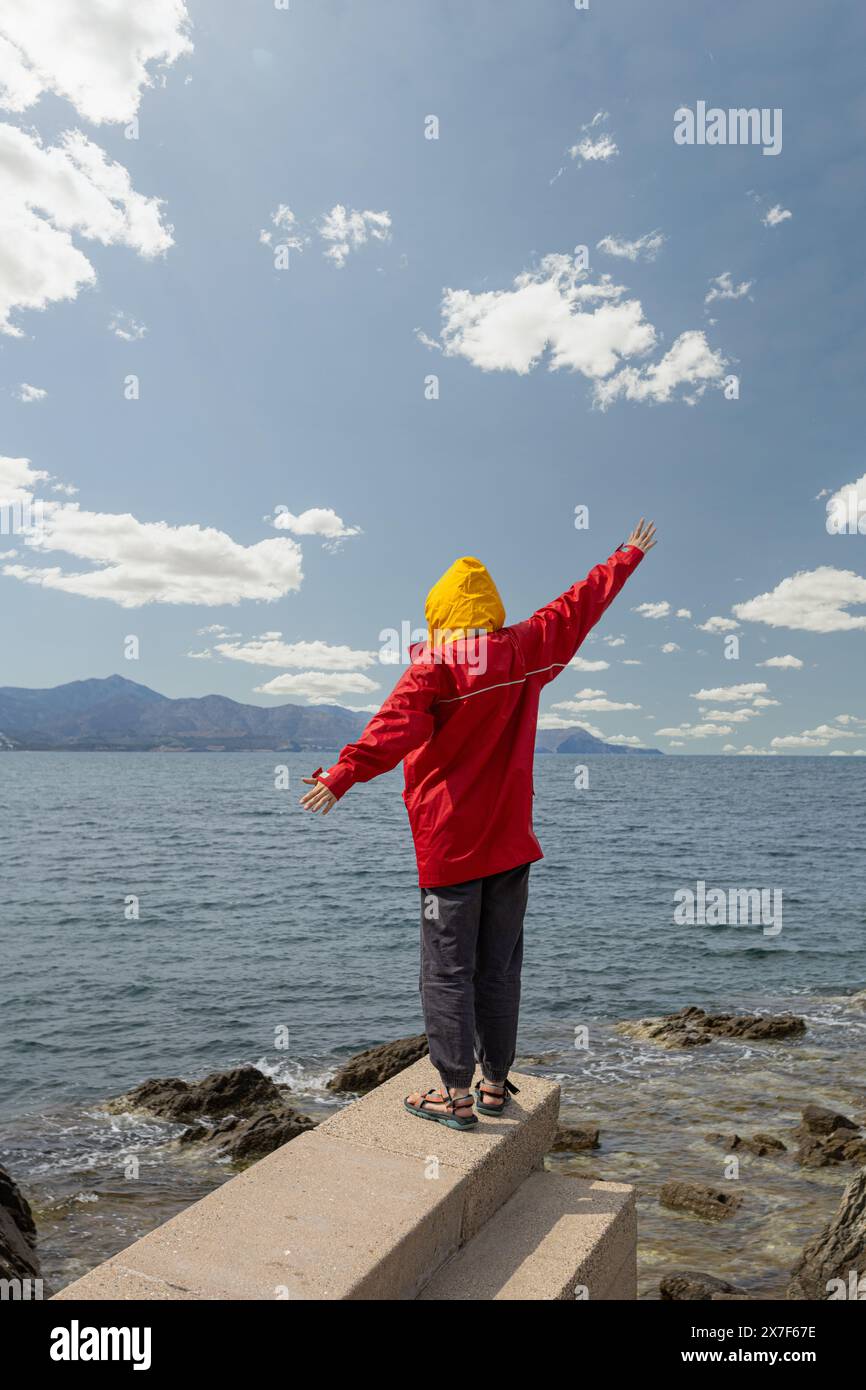 person in front of the sea with a storm overhead with large gray clouds, weather change red jacket with yellow cap, arms raised in protest Stock Photo