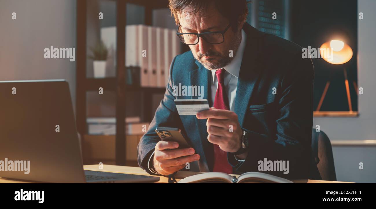 Businessman completing online credit card purchase with mobile phone app in business office late at night, selective focus Stock Photo