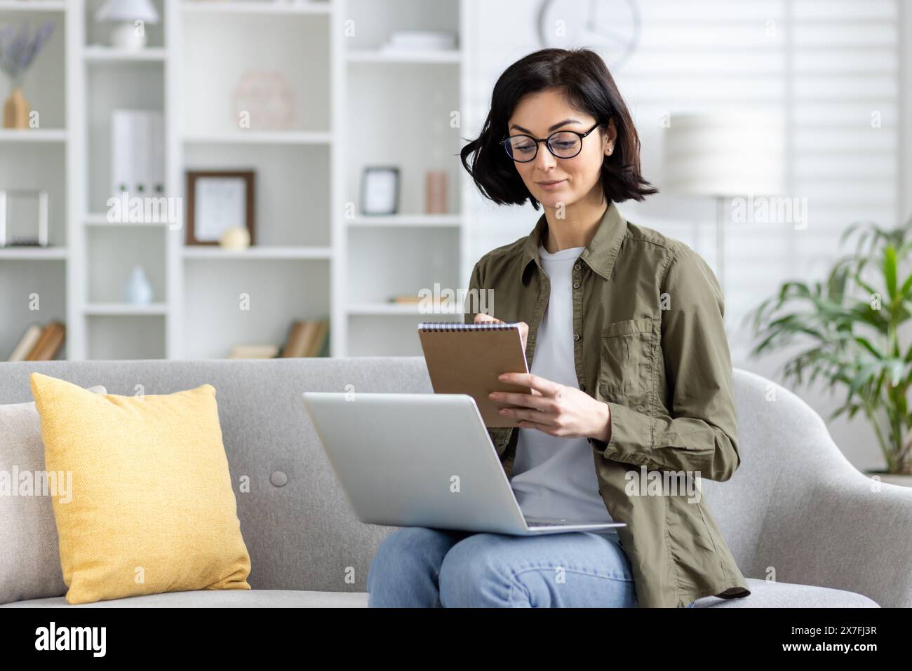 Young concentrated and serious woman sitting on the sofa at home and studying remotely, working on a laptop and making notes in a notebook. Stock Photo