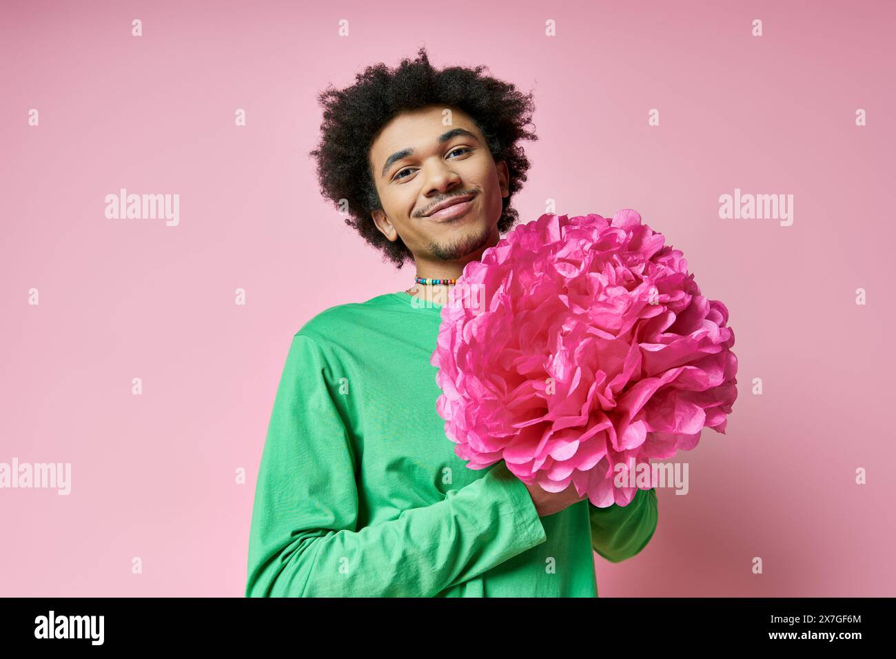 A cheerful, curly-haired African American man in casual wear holds a large pink flower against a pink background. Stock Photo