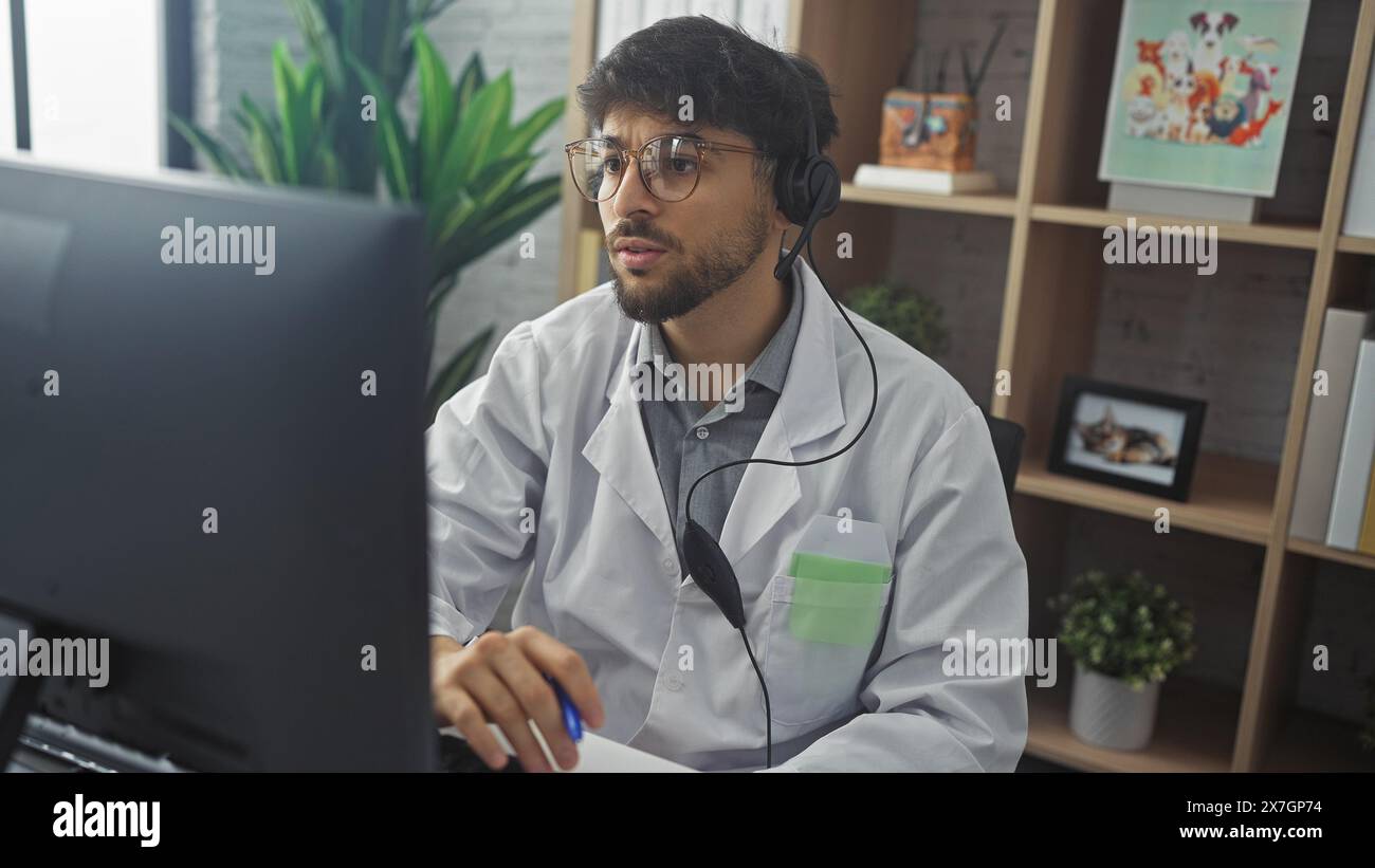 Handsome arab male doctor in headset working at clinic office with computer, paperwork, and indoor plants. Stock Photo