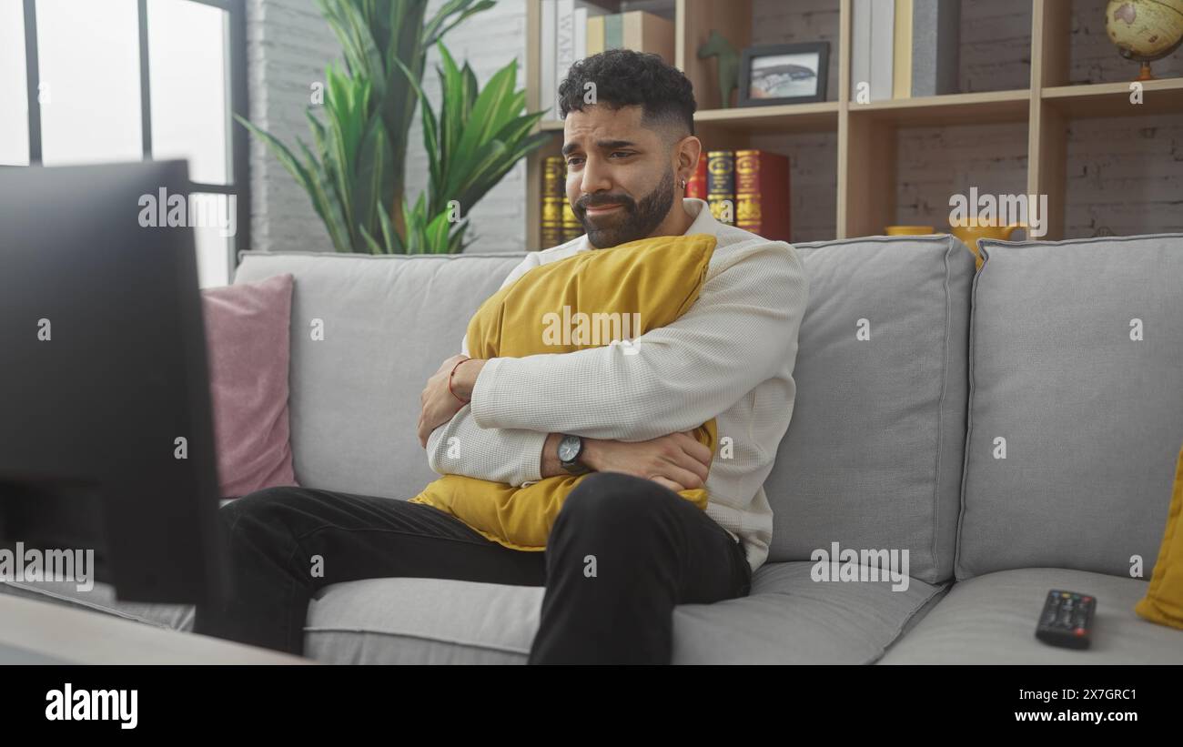 Handsome hispanic man with beard sitting on a gray sofa looking sad while watching tv in a cozy living room. Stock Photo