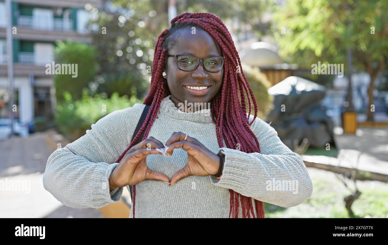 A cheerful african woman with braids forming a heart sign outdoors in ...