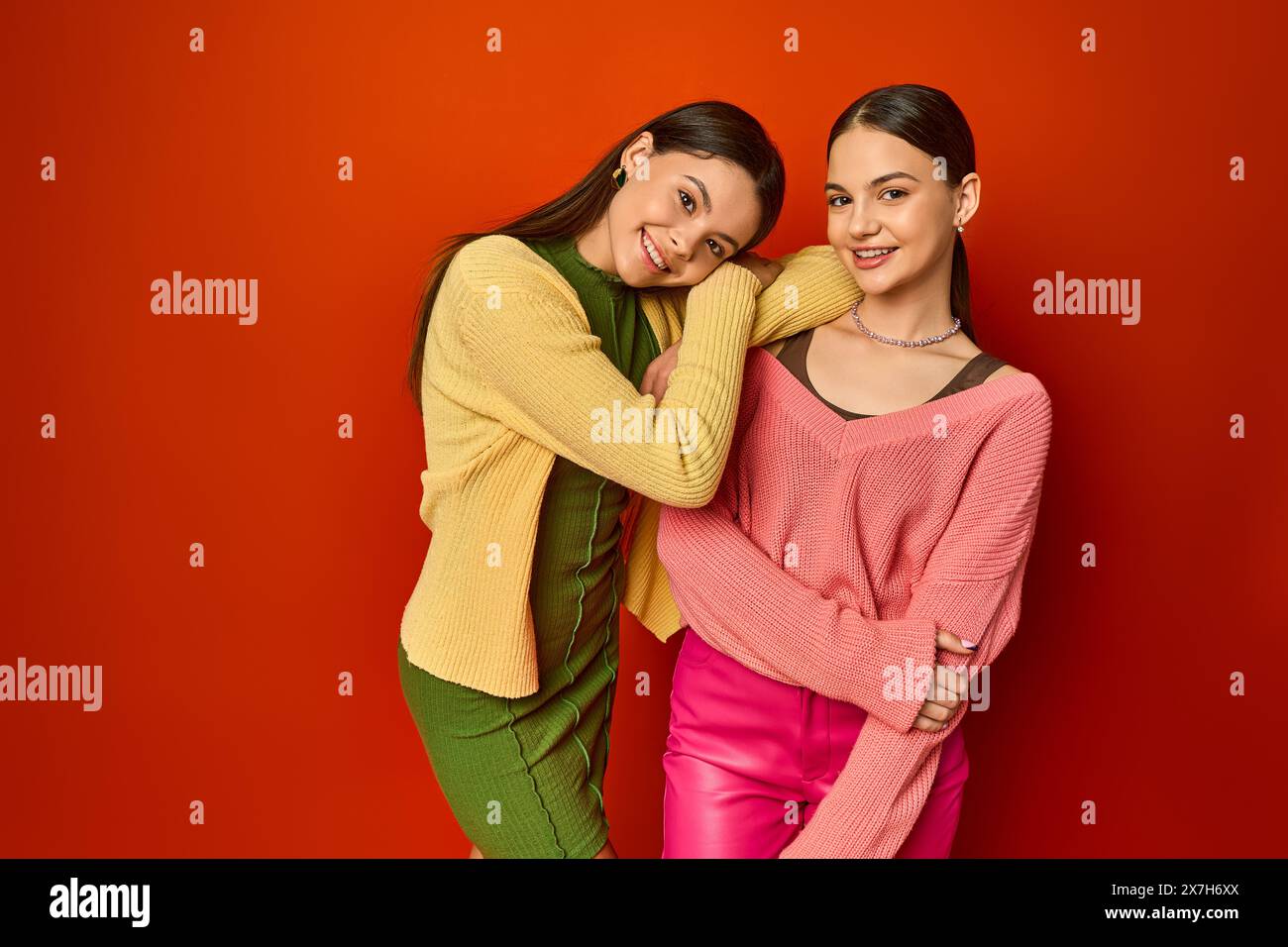 Two pretty brunettes, teenage friends, standing together in front of a vibrant red wall in a studio setting. Stock Photo