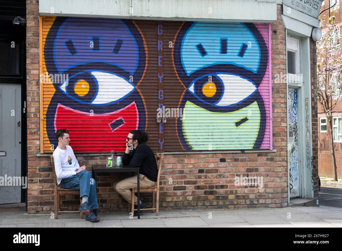 Hackney, Goldsmiths Row. Two young men sit at a table with drinks under a colourful shop blind Stock Photo