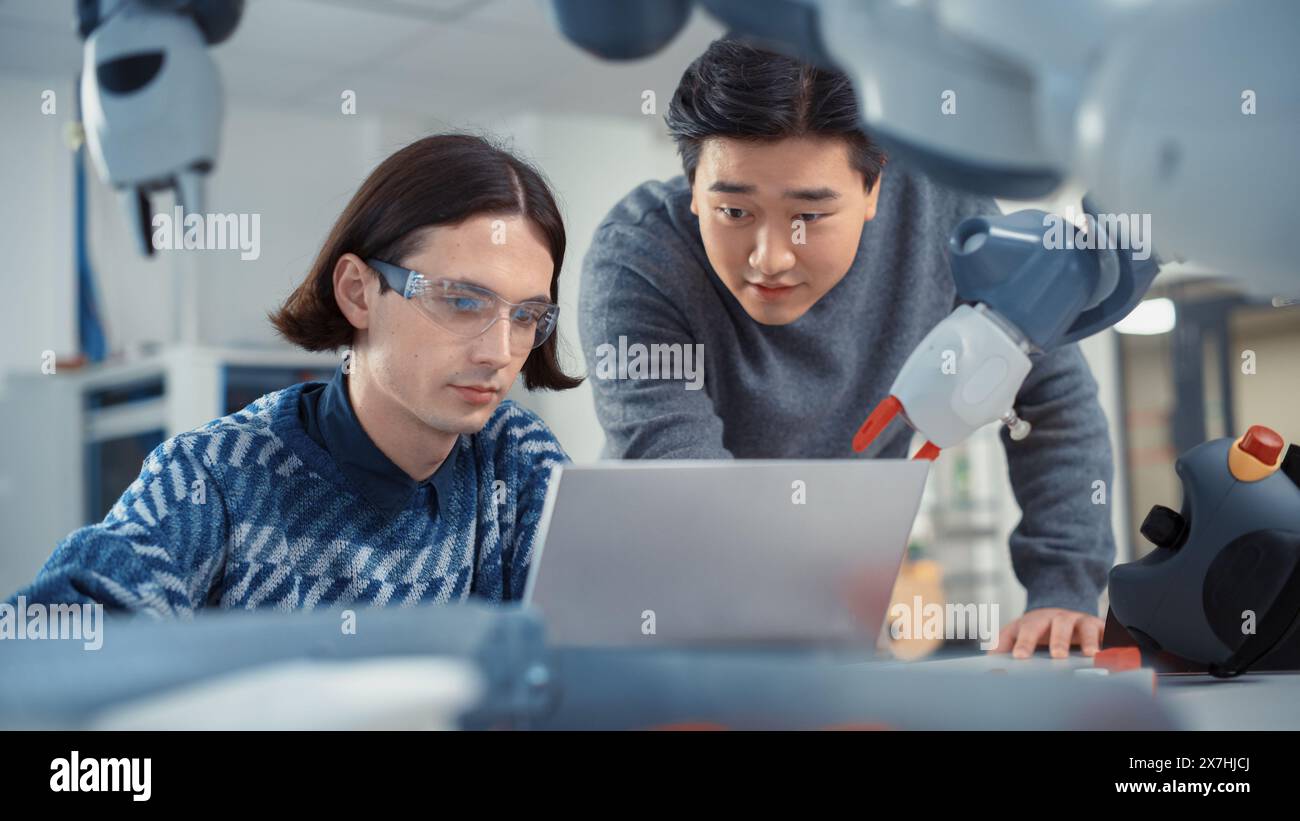 Two Diverse Developers Brainstorming and Coding On Laptop. Asian Male Press Button on Control Panel. Colleagues Building, Configuring and Testing Robots. Automation Technologies Concept. Stock Photo
