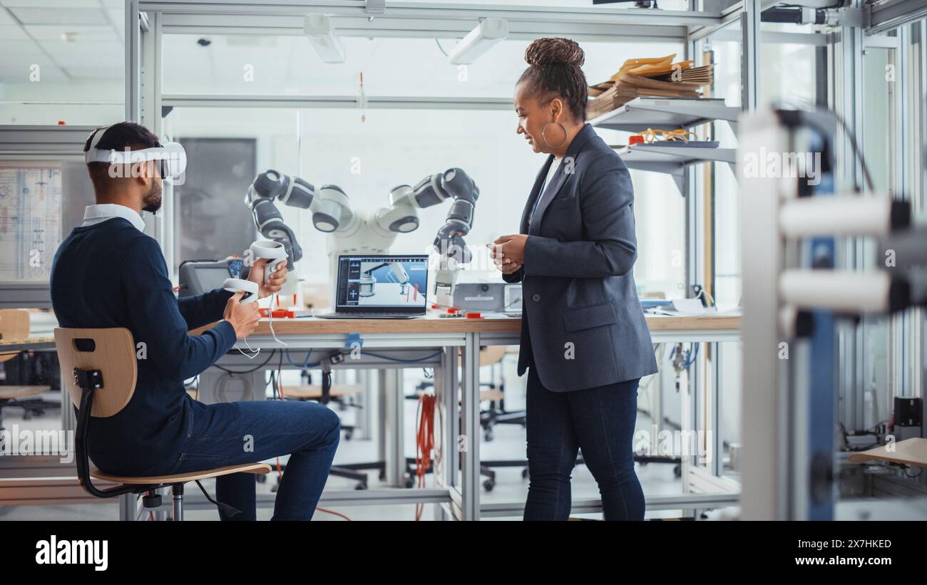Black Senior Engineer Talks to Young Male Computer Scientist Who is Wearing Virtual Reality Headset and Uses Controllers to Remotely Manipulate Robotic Hand. High Tech Research Laboratory Concept Stock Photo