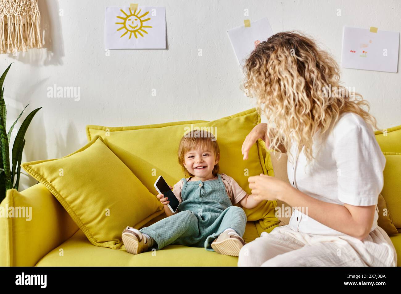 A curly-haired mother lovingly combs her toddler daughters hair while sitting on a cozy yellow couch at home. Stock Photo