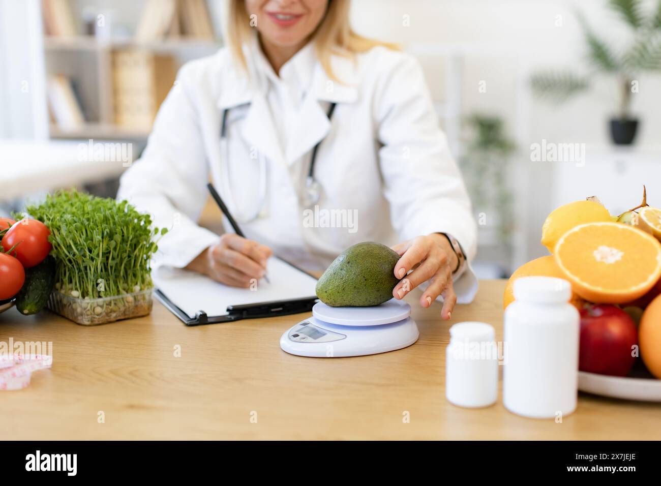 Close up of hands of mature female nutritionist making healthy eating plan and calculating calorie content of avocado using weight. Happy female writi Stock Photo