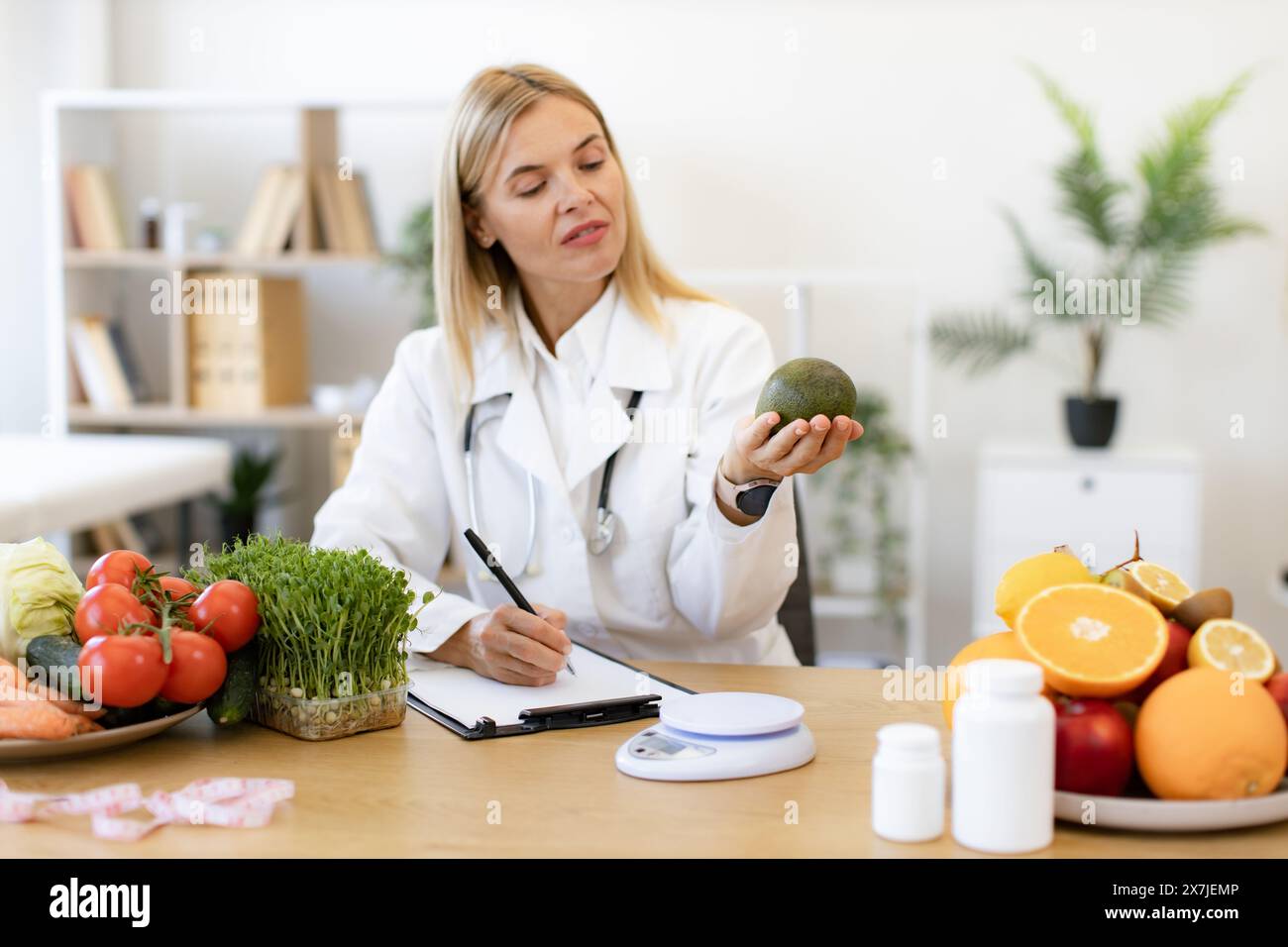 Mature Caucasian female nutritionist making healthy eating plan and calculating calorie content of avocado using weight. Happy female writing prescrip Stock Photo