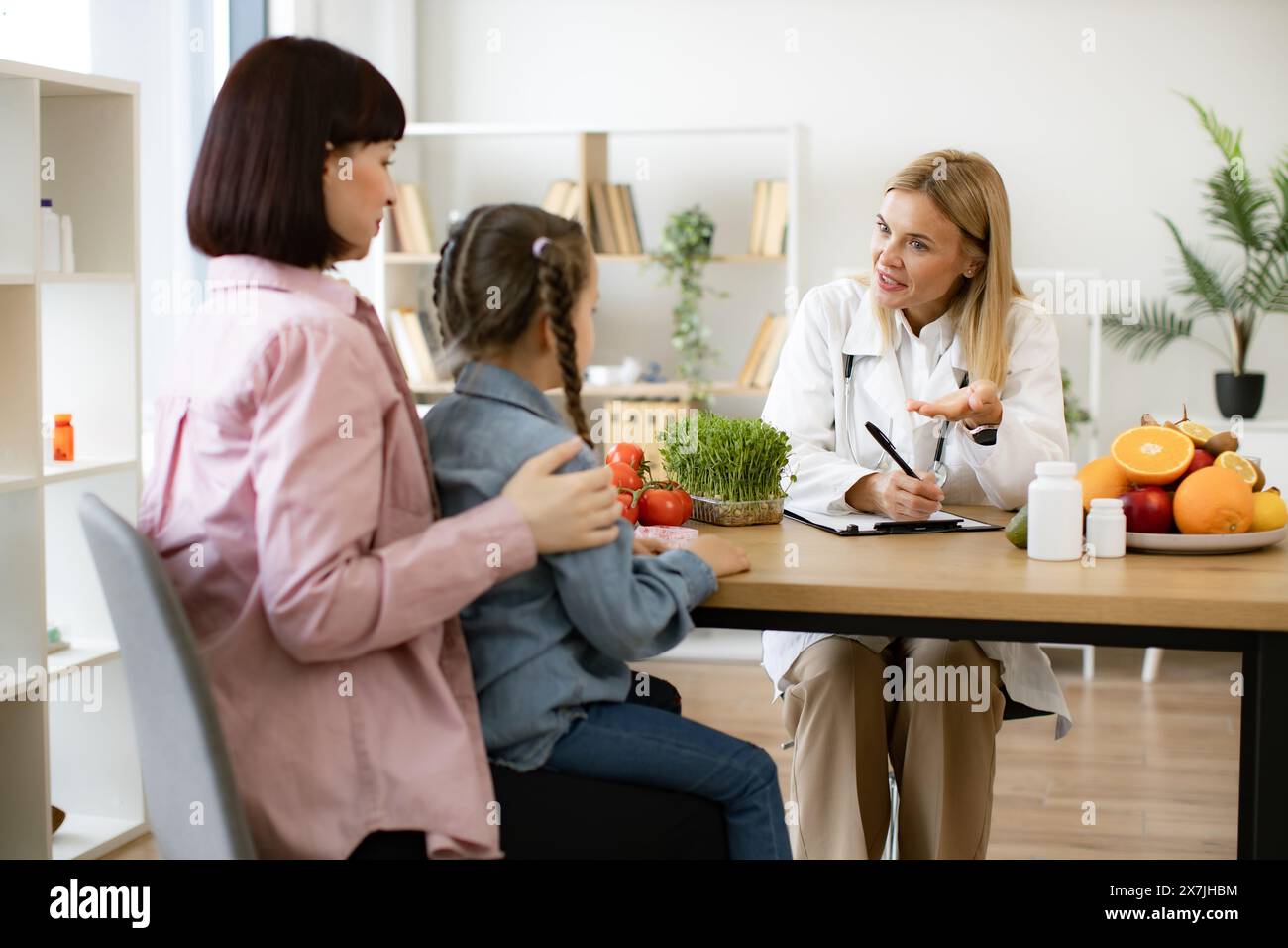 Mature Caucasian female doctor talking with child girl sitting on her mom knees and writing diet plan. Experienced female dietitian prepares course of Stock Photo