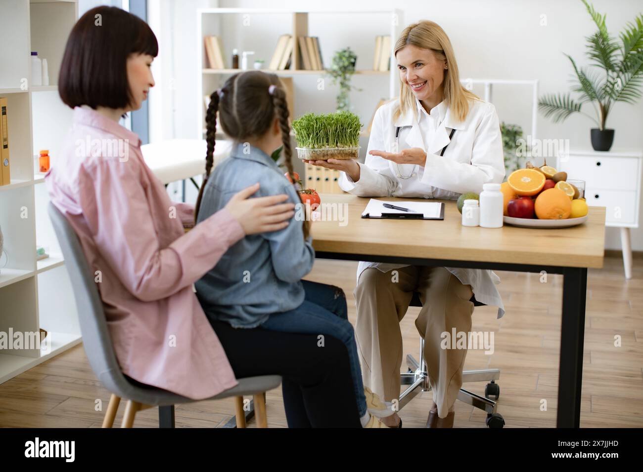 Little girl sitting on mother lap at appointment with mature female nutritionist making healthy eating plan. Beautiful female doctor holding microgree Stock Photo