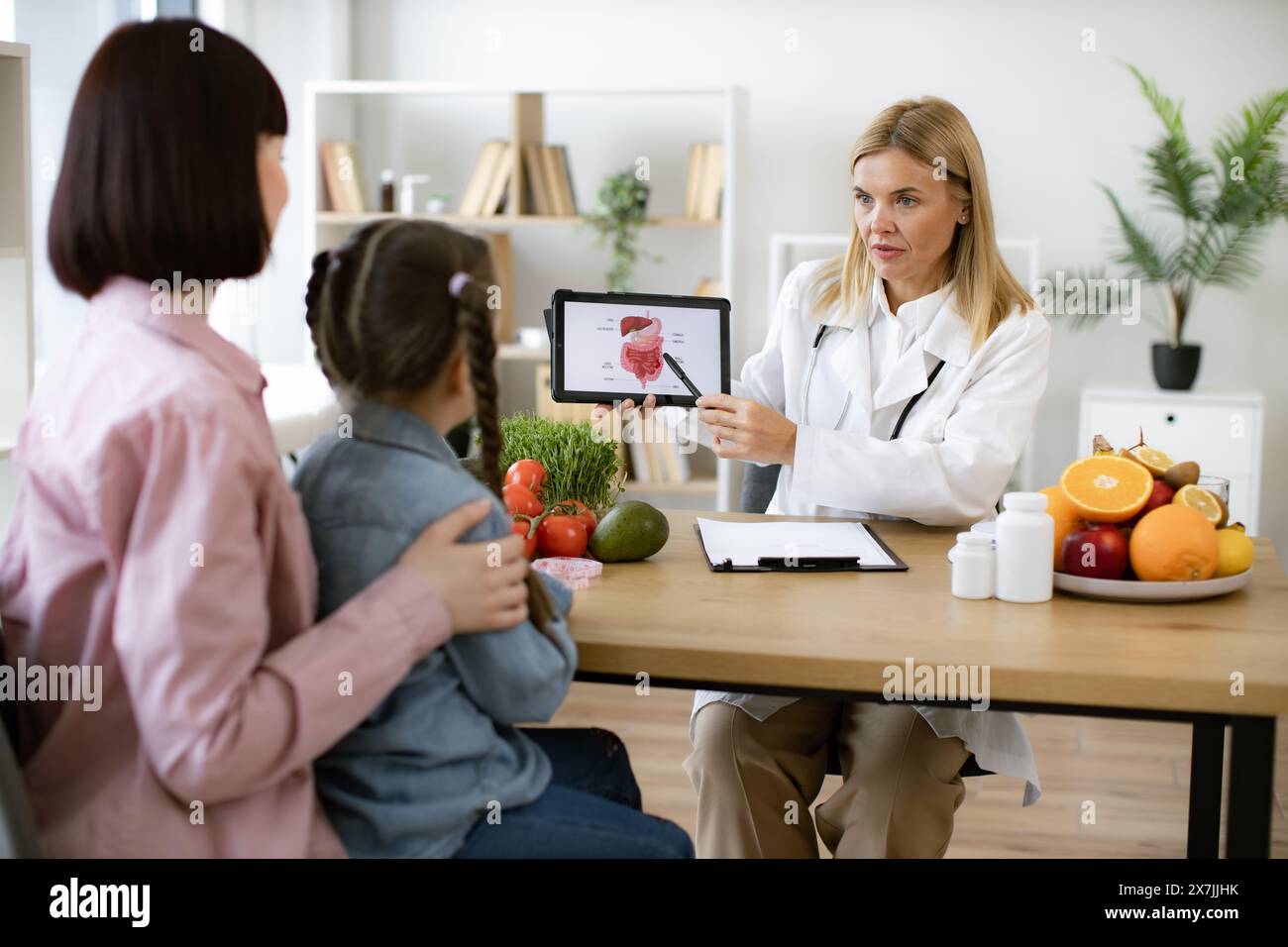 Mature experienced female doctor showing tablet with gastrointestinal tract image for treatment planning to little female child. Mother with little gi Stock Photo