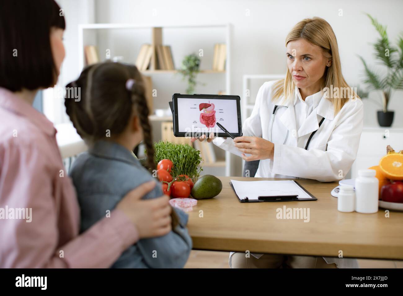 Mature experienced female doctor showing tablet with gastrointestinal tract image for treatment planning to little female child. Mother with little gi Stock Photo
