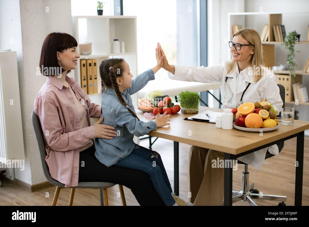 Little girl sits on mothers lap and gives high five to mature positive female nutritionist after successful treatment for indigestion. Concept of comp Stock Photo