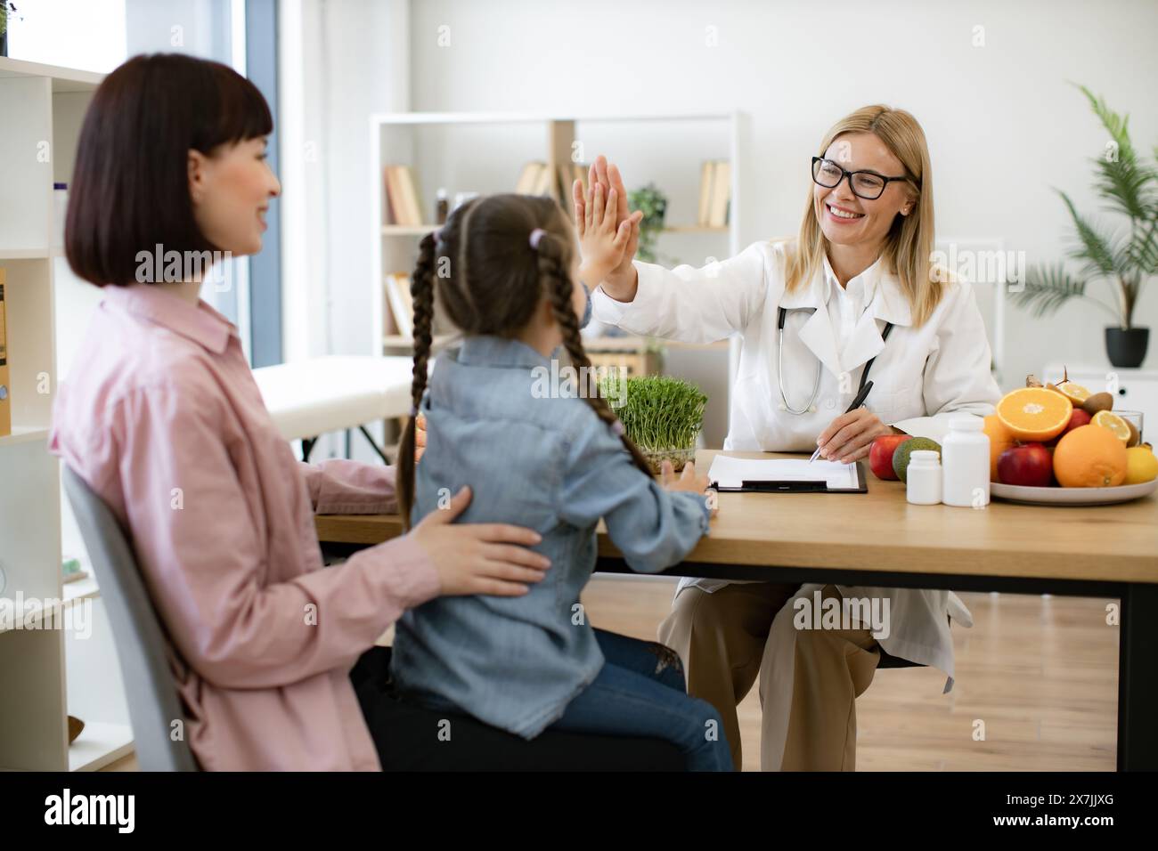 Little girl sits on mothers lap and gives high five to mature positive female nutritionist after successful treatment for indigestion. Concept of comp Stock Photo