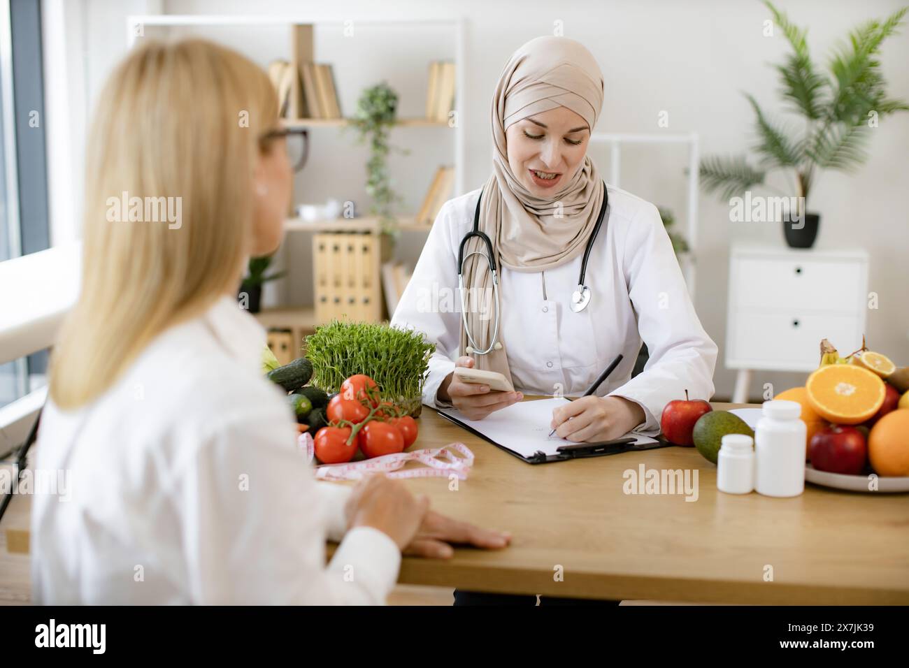 Experienced female dietitian preparer course of healthy nutrition for adult woman. Muslim female doctor sitting at table writing diet plan talking wit Stock Photo