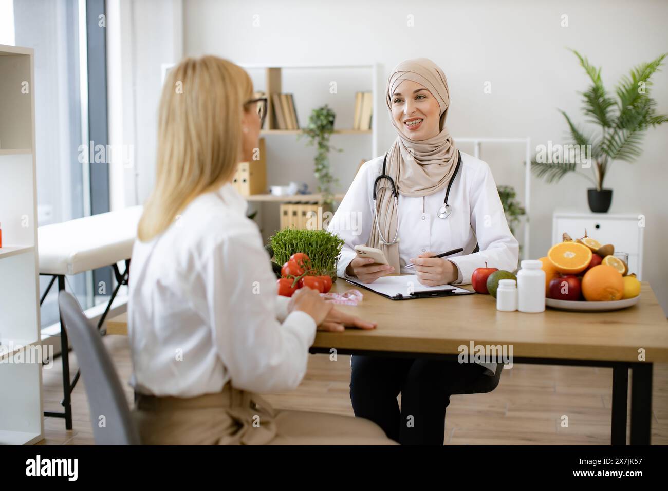 Muslim female doctor sitting at table writing diet plan talking with mature patient lady. Experienced female dietitian preparer course of healthy nutr Stock Photo
