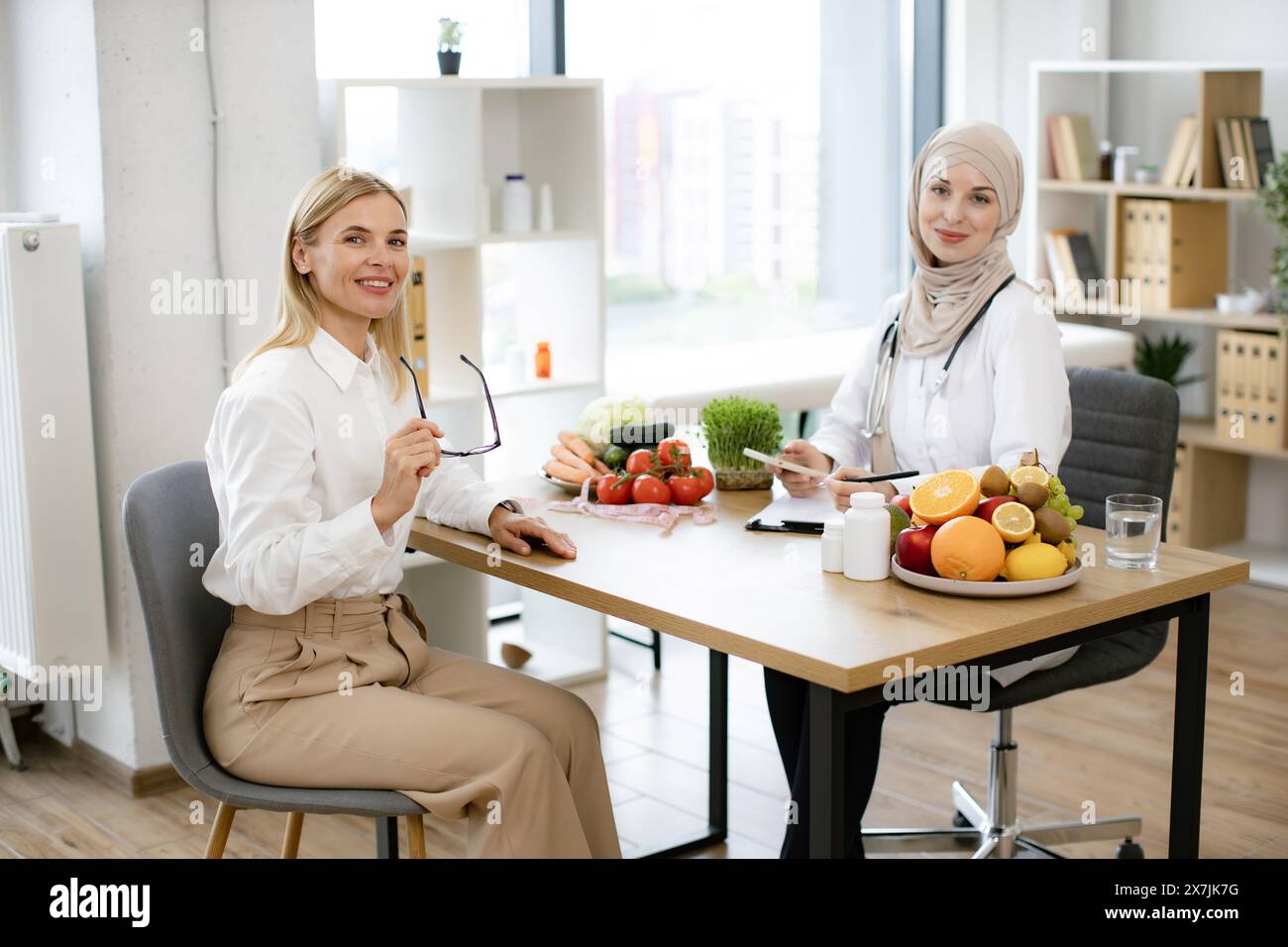 Young Arab female doctor making healthy eating plan. Muslim nutritionist in hijab writes electronic prescription to female patient using phone while s Stock Photo