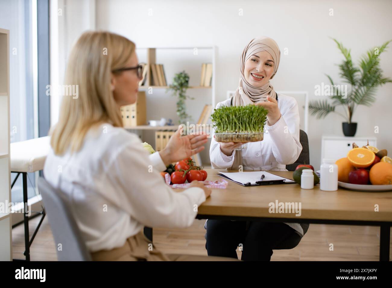 Mature lady sitting on appointment with muslim female nutritionist making healthy eating plan. Beautiful female doctor holding microgreen while adding Stock Photo