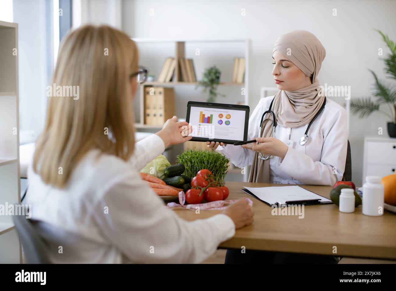 Professional nutritionist tells patient about benefits of healthy diet while holding tablet with graphs in hands. Arab female doctor makes treatment p Stock Photo