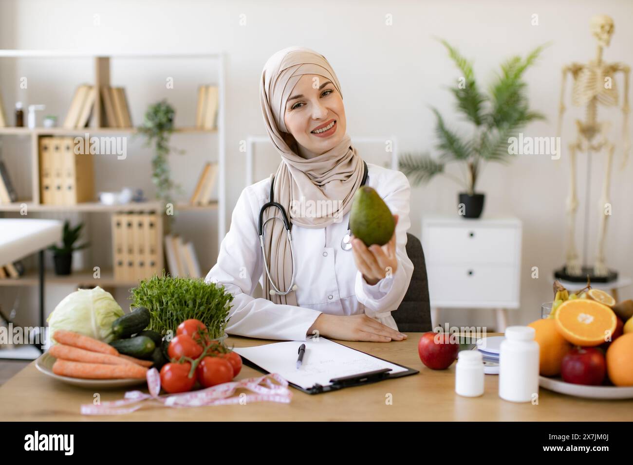 Arabian female nutritionist making healthy eating plan and calculating calorie content of avocado using weight. Happy female writing prescription for Stock Photo