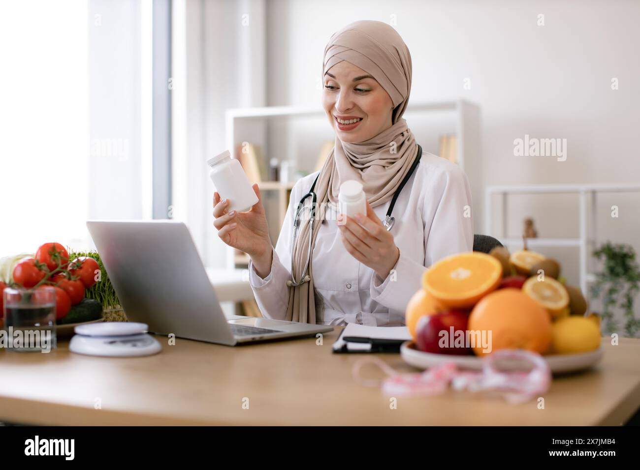 Muslim doctor in hijab distantly describes the treatment plan. Positive arabian nutritionist woman holding pills or vitamins in hands during online co Stock Photo