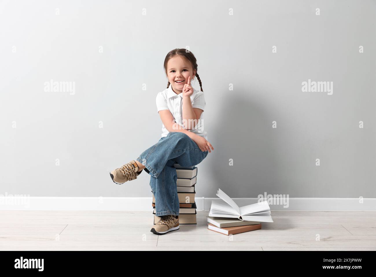 Cute little girl sitting on stack of books near light grey wall Stock ...