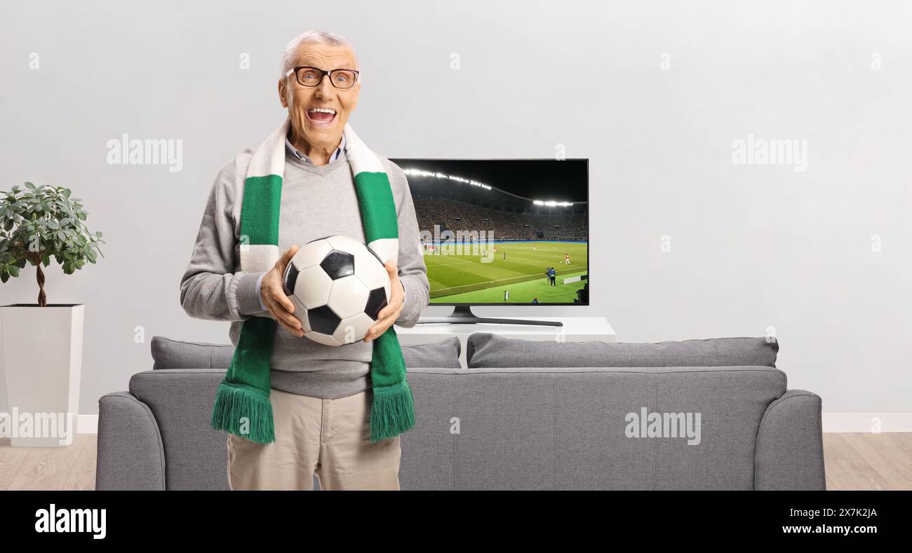 Excited elderly man in front of a sofa and tv holding a football and wearing a scarf at home Stock Photo