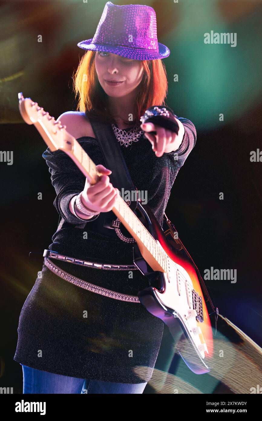 joyful young woman with red hair, in a glittering black ensemble and sequined hat, skillfully plays her electric guitar, stage lights casting colorful Stock Photo