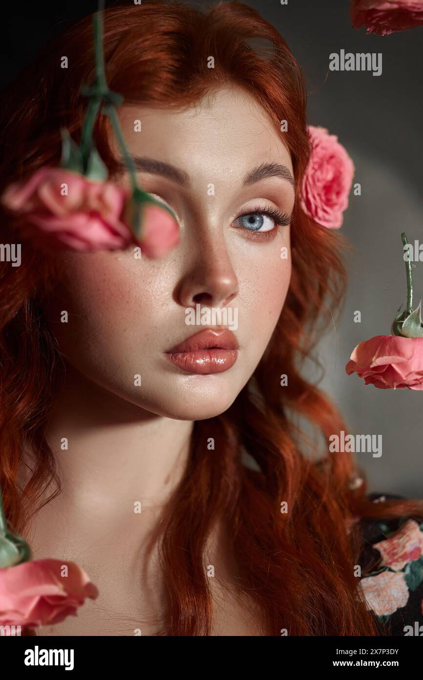 Red-haired woman with striking blue eyes looks away, surrounded by delicate pink roses Stock Photo