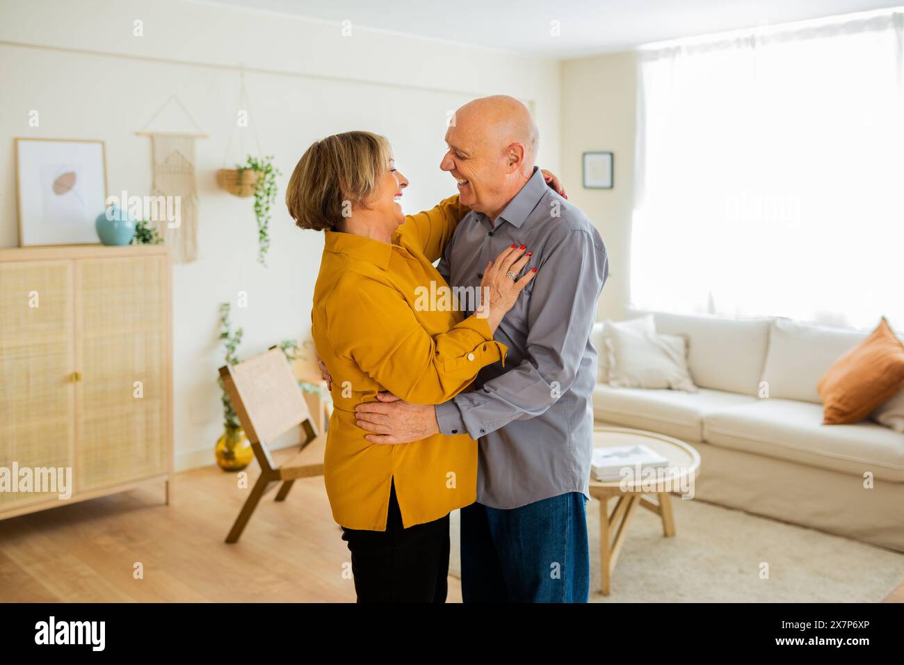 Senior couple dancing together in their living room, smiling and looking into each other's eyes. Couple enjoying a tender moment at home. Stock Photo