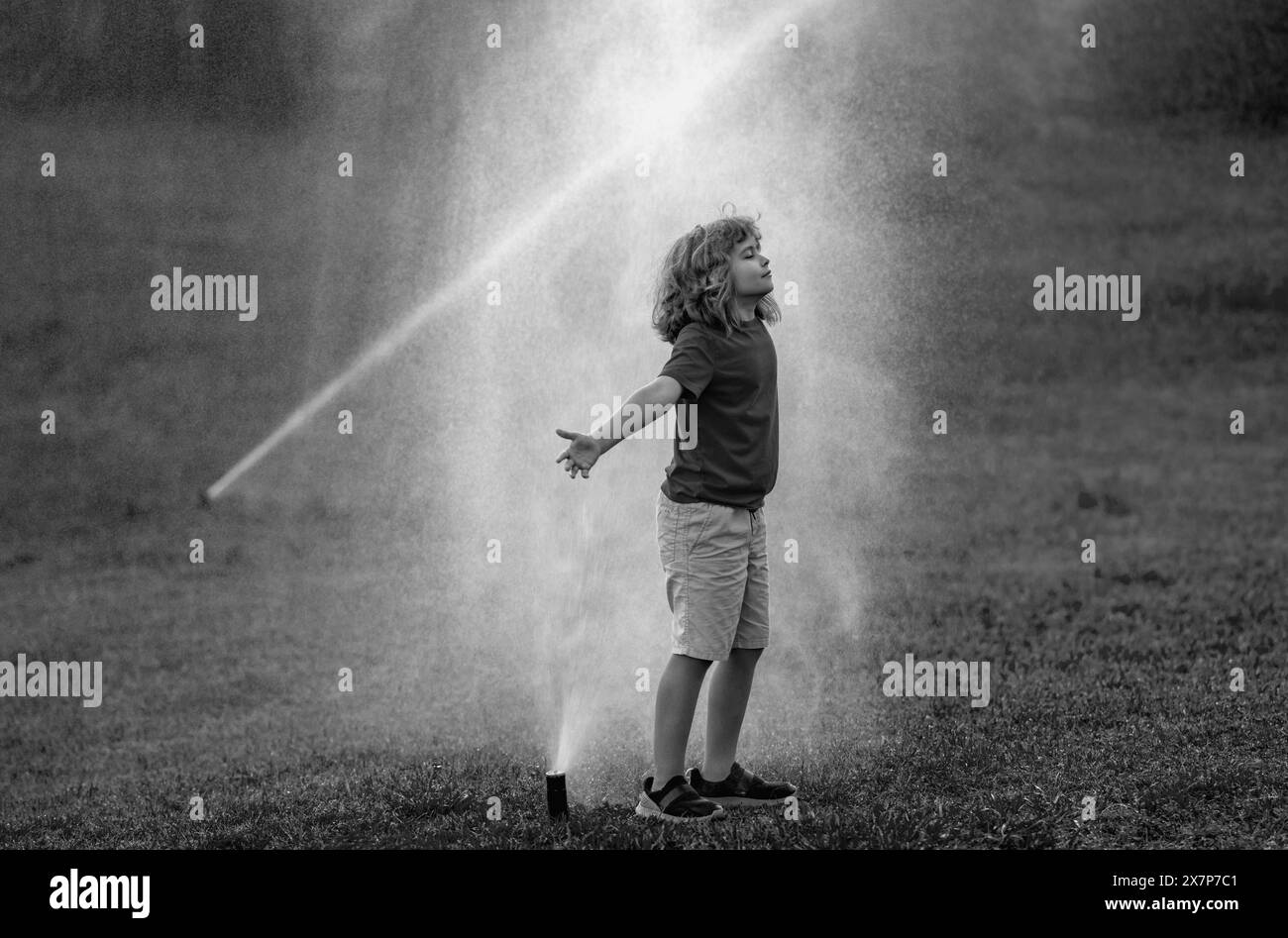 Child play in summer garden. Grass watering. Automatic sprinkler irrigation system in a green park watering lawn. Sprinkler watering. Child gardening Stock Photo