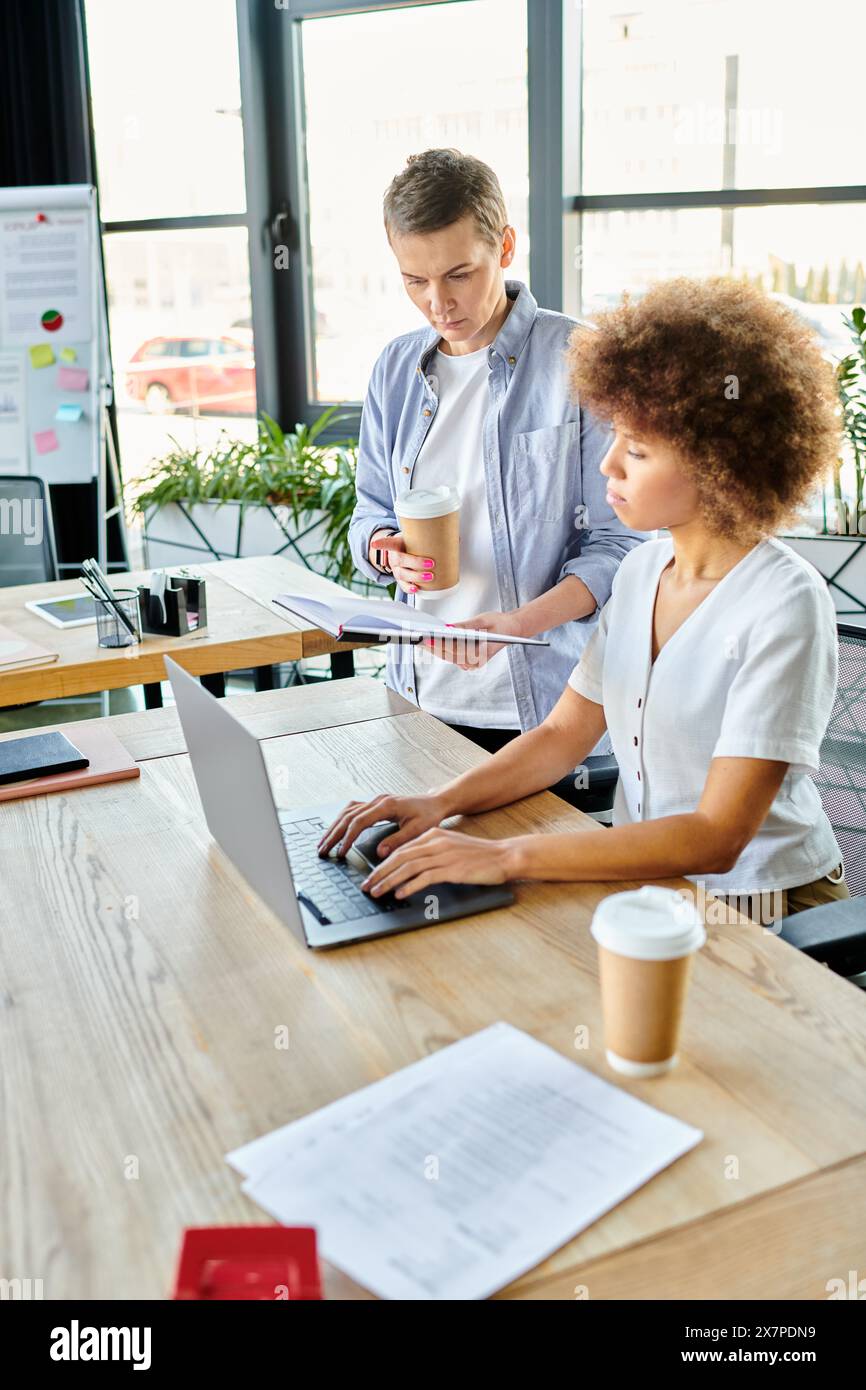 Two diverse women collaborating on a laptop at a table in a modern workspace. Stock Photo