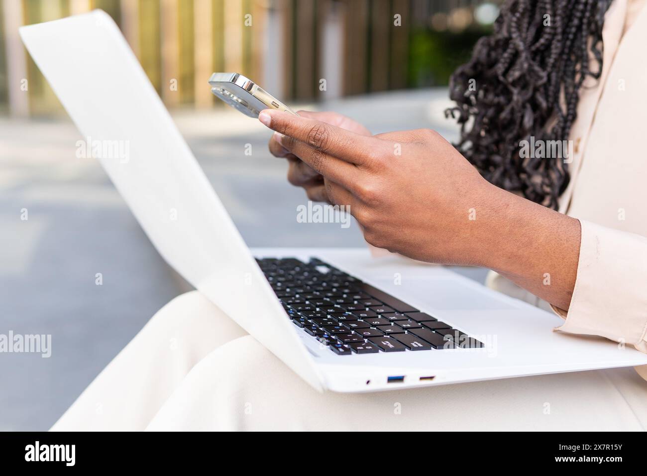 Close-up of an African American businesswoman using her smartphone while typing on a laptop, focusing on multitasking with technology outdoors Stock Photo