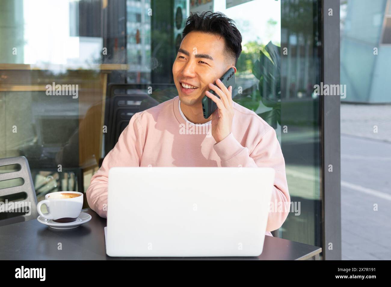 Smiling Asian man in casual attire having a conversation on his smartphone while working on a laptop in a cafe setting, with a cup of coffee on the ta Stock Photo
