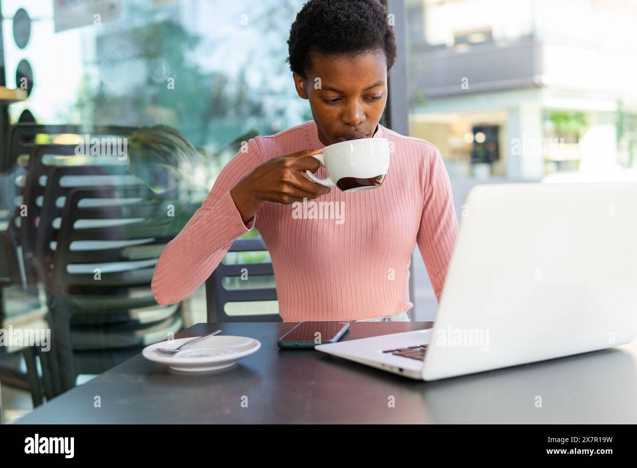 An African woman is focused on her laptop screen while sipping coffee at a cafe, reflecting productivity and a casual work environment. Stock Photo