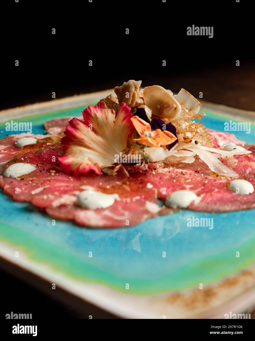 Elegant presentation of a beef carpaccio, adorned with edible flowers, mushrooms, and dollops of sauce on a vibrant ceramic plate Ideal for fine dinin Stock Photo