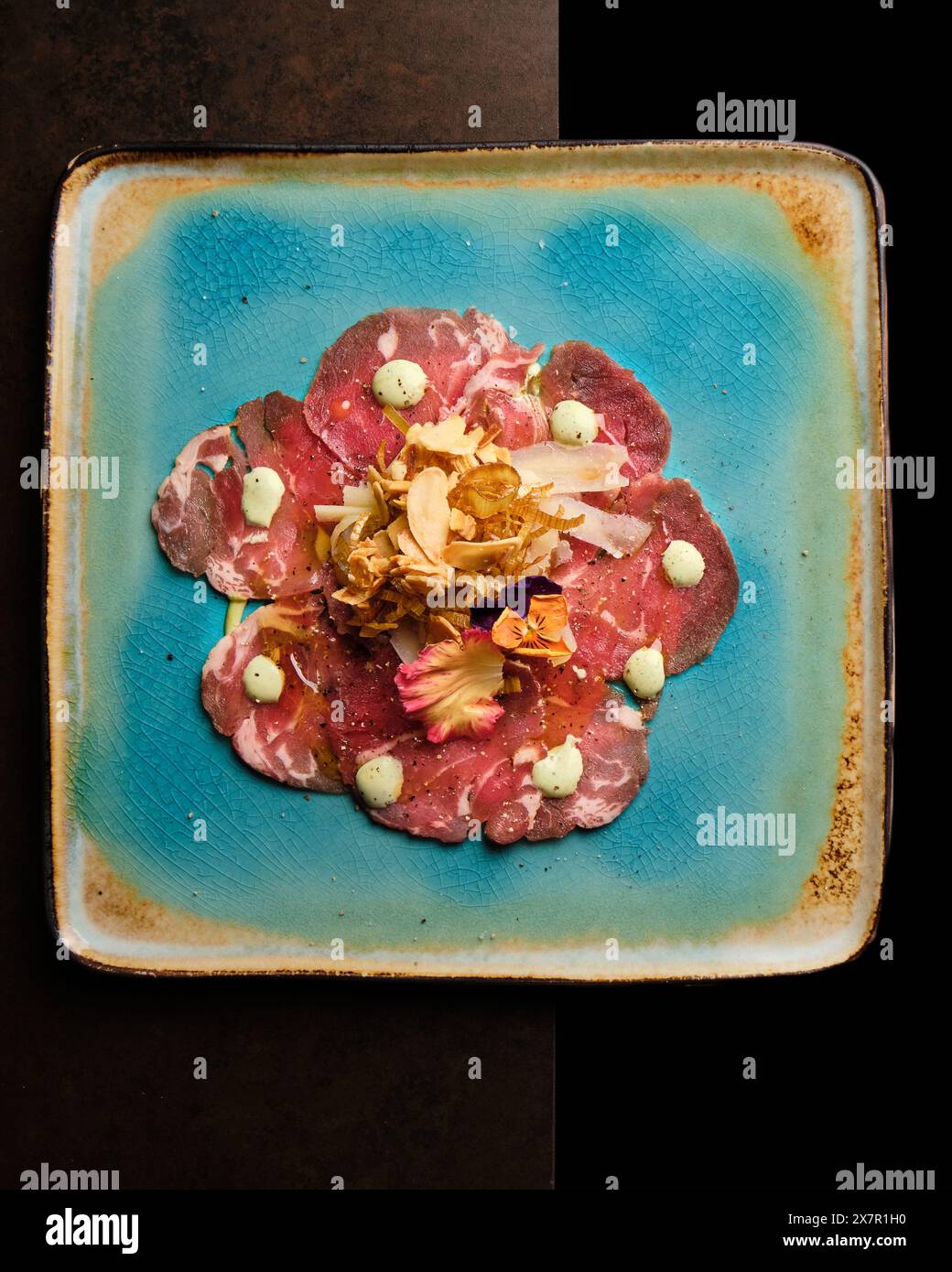 Artistic presentation of beef carpaccio adorned with dollops of sauce, edible flowers, and crispy toppings, elegantly served on a rustic blue plate ag Stock Photo