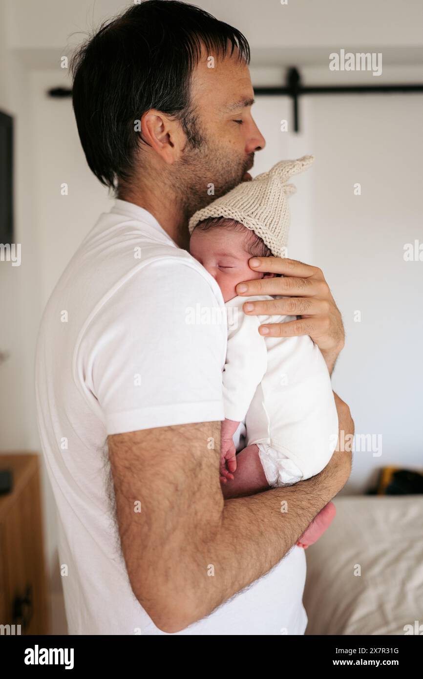 A father lovingly holds his newborn baby, who wears a knit cap, in a ...