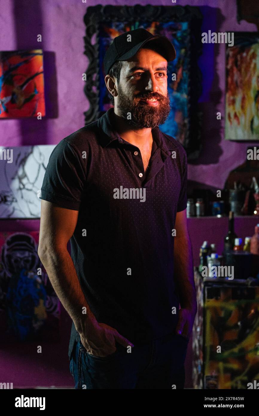 A Latin American artist stands thoughtfully with hands in his pockets in his studio, surrounded by vibrant paintings reflecting his heritage and the c Stock Photo