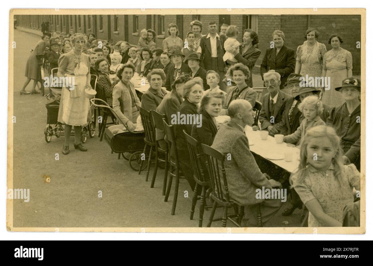 Original, evocative WW2 era postcard of end of World War 2 street party celebration, women and children and older men, taking tea outside on a long table in their urban street, lots of characters, a cheeky young girl looks into the camera. A lively scene.  A typical British street party with many cups of tea. Mainly party for older women and a few men. Vintage Devon. Photo dated August 1945, Salisbury Street, Exmouth, Devon, England, U.K. Stock Photo