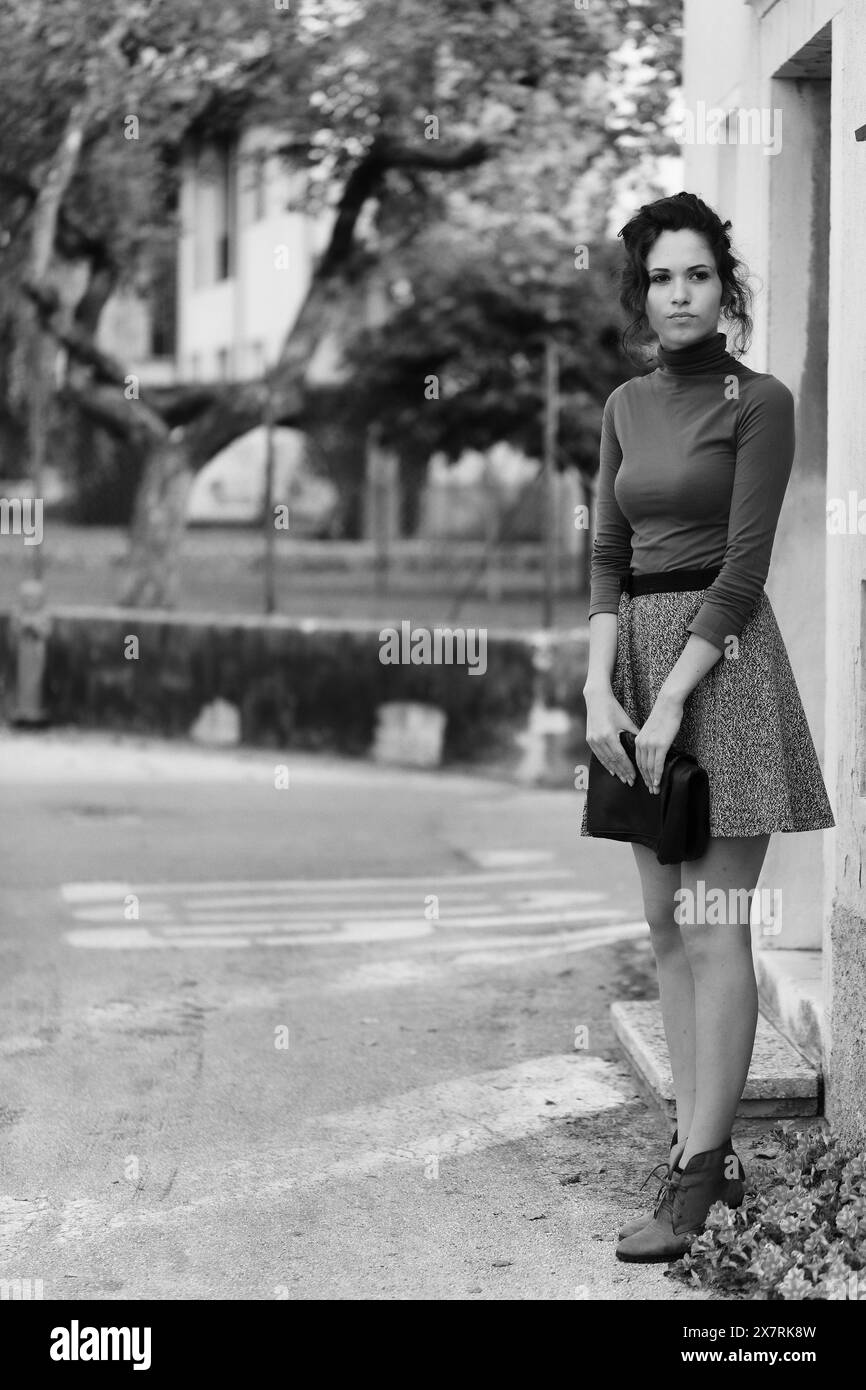 young woman stands near a building, appearing to wait for someone. She wears a brown turtleneck and grey skirt, holding a clutch. The black and white Stock Photo