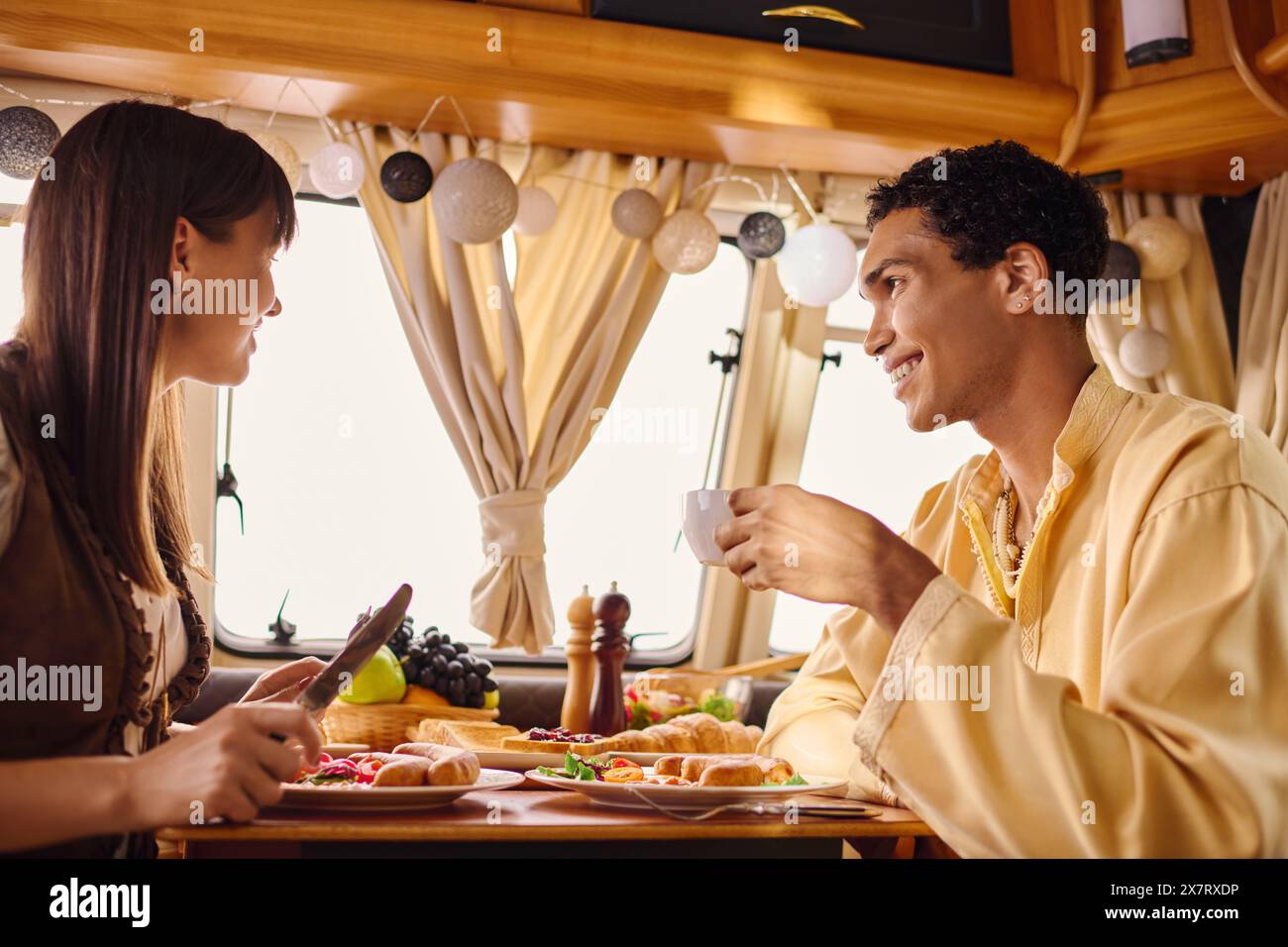 An interracial man and woman sitting at a table, enjoying a delicious meal together in a camper van on their romantic getaway. Stock Photo