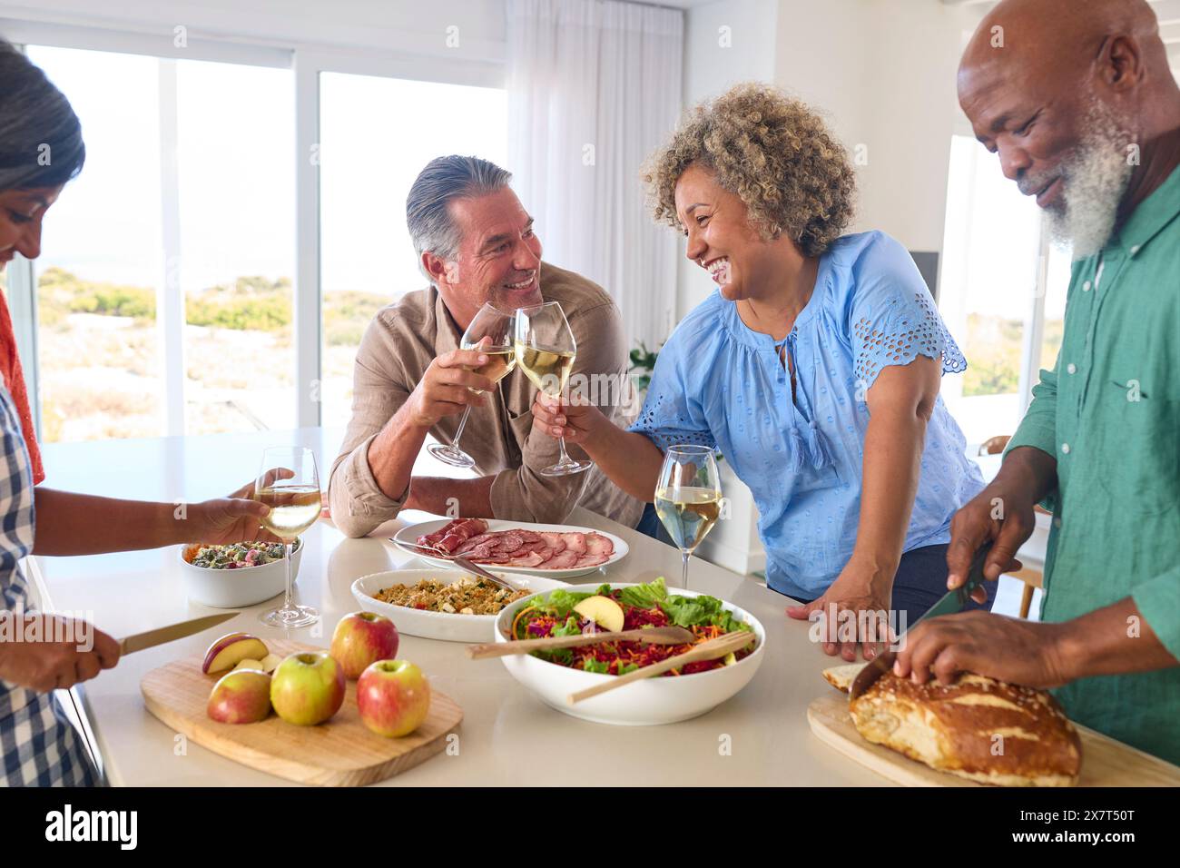 Group Of Smiling Mature Friends At Home Relaxing Preparing Food For Lunch With Wine Together Stock Photo